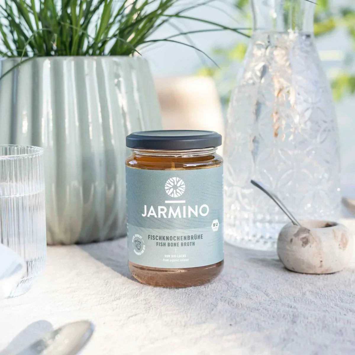 Jarmino Fischknochenbrühe 350ml – Glasflasche auf Sand, dekorativ arrangiert, natürliche Atmosphäre